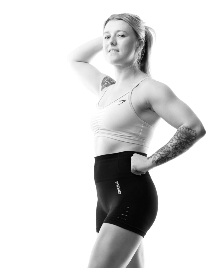 A woman in athletic wear poses against a white background, hand behind head, looking to the side.