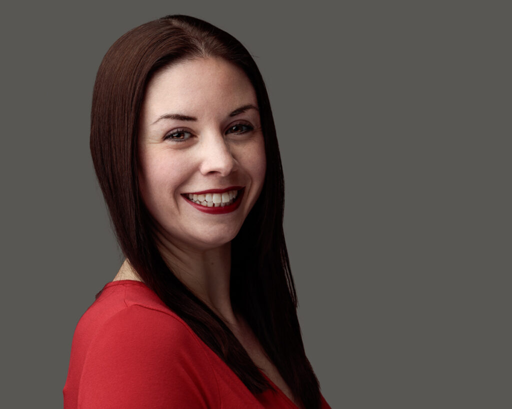 Woman with long dark hair wearing a red top, smiling at the camera against a neutral grey background.