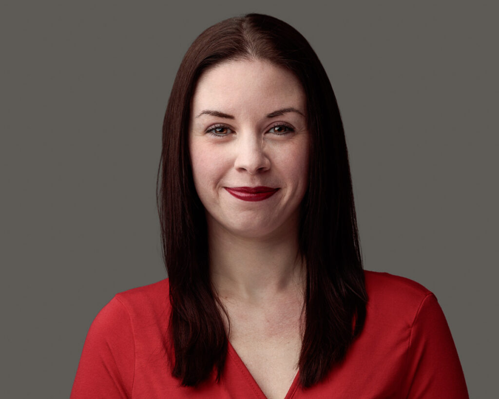 A woman with long dark red hair and red lipstick, wearing a bright red top, against a neutral grey background.