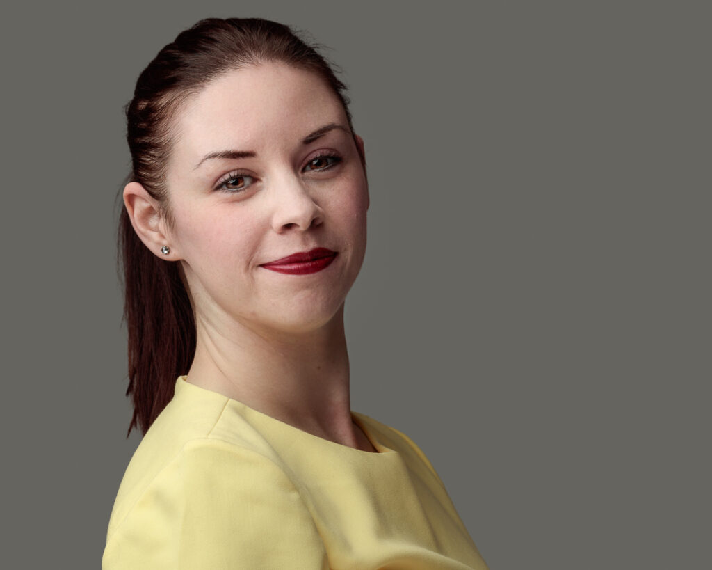 A woman with dark reddish-brown hair wearing a yellow top, looking over her shoulder against a plain grey background.
