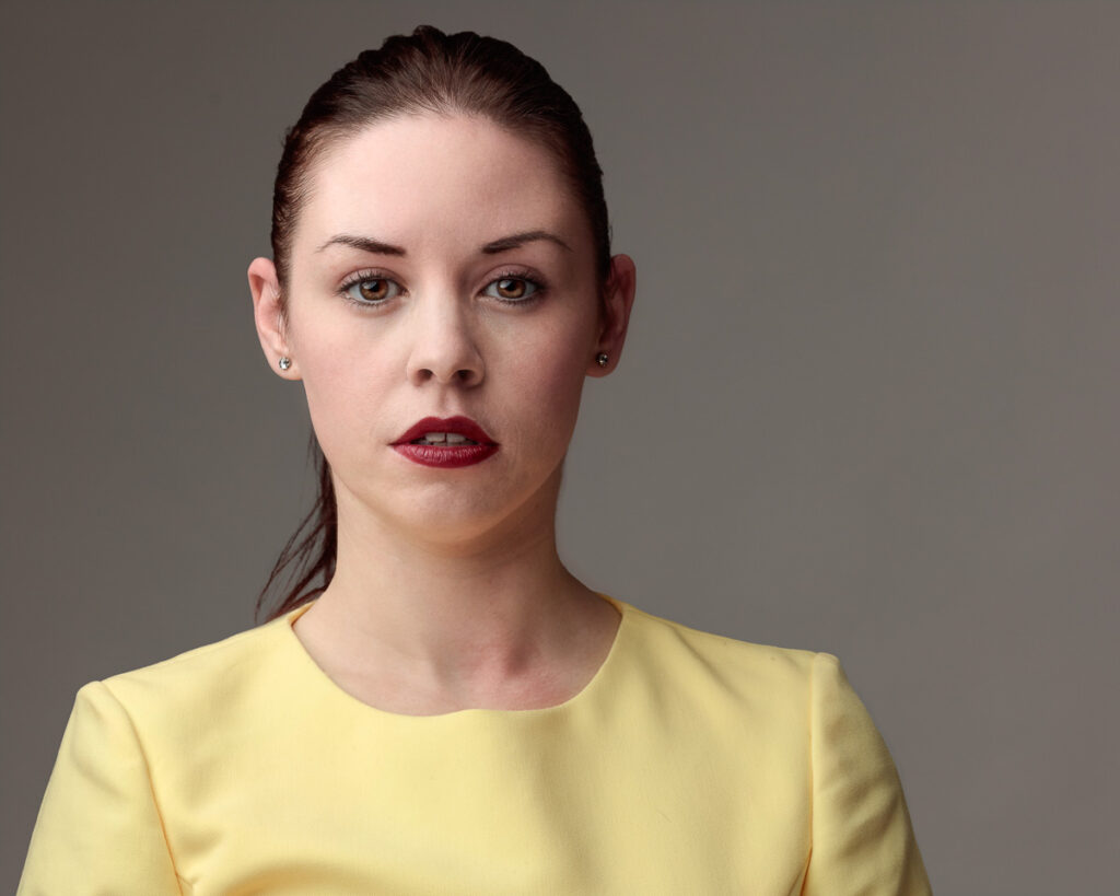 Woman with dark hair pulled back, wearing a yellow top and red lipstick, against a grey background, looking directly ahead with a neutral expression.