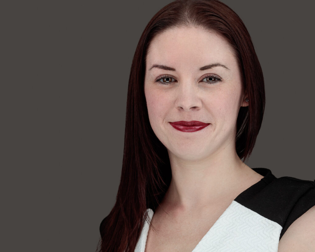 A woman with long dark reddish-brown hair and burgundy lipstick looks downward against a grey background.