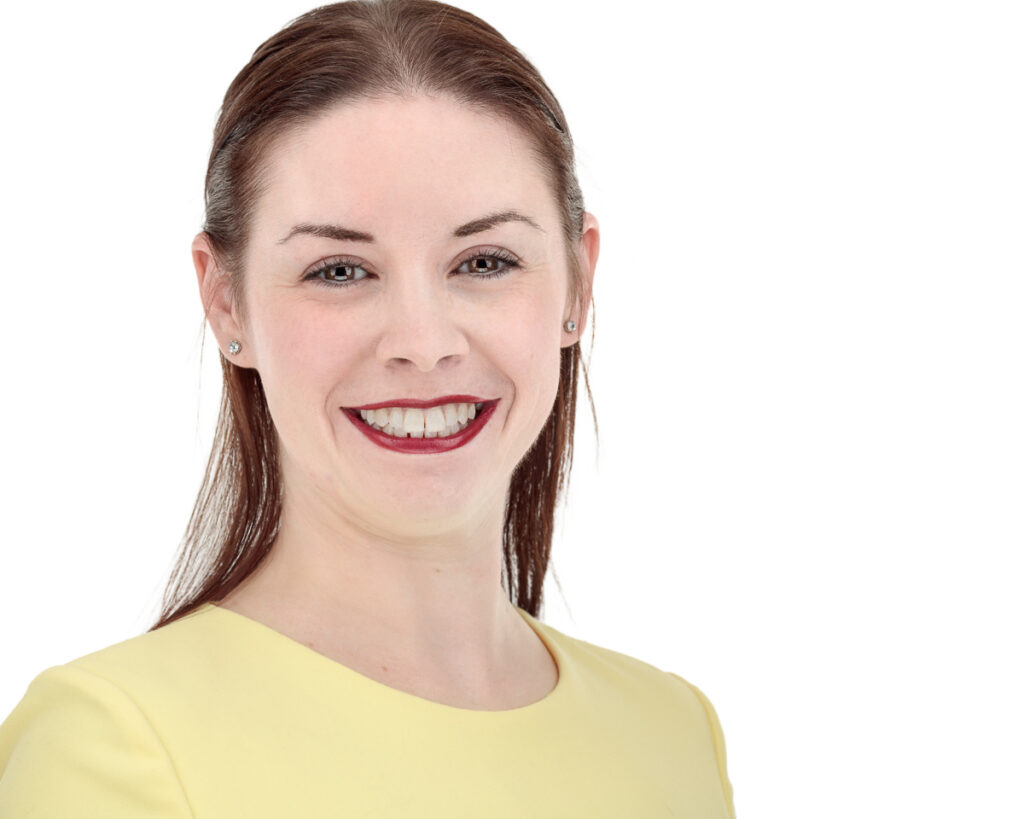 A woman with reddish-brown hair wearing a yellow top and burgundy lipstick smiles at the camera against a white background.