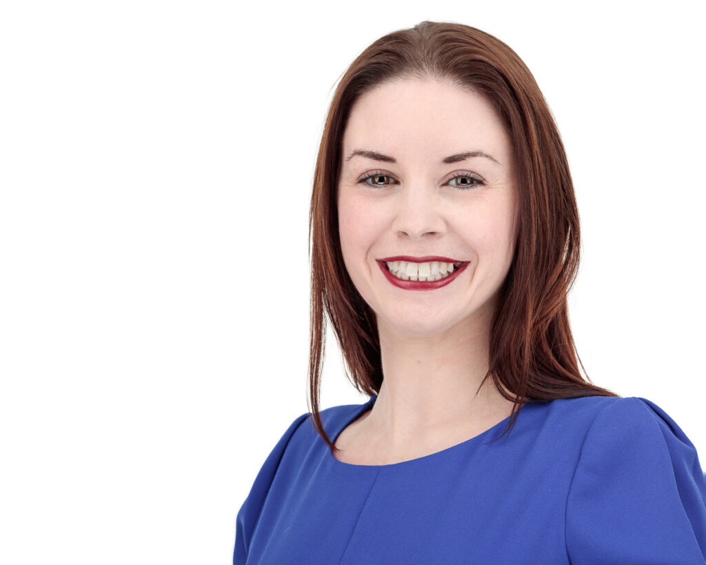 A woman with shoulder-length reddish-brown hair wearing a blue top, smiling at the camera against a white background.
