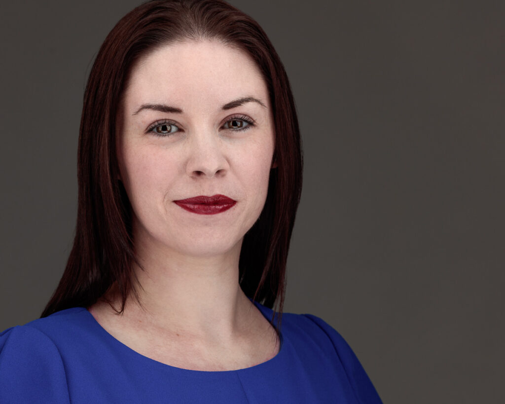 A woman with dark reddish-brown hair and burgundy lipstick wears a blue top against a grey background.