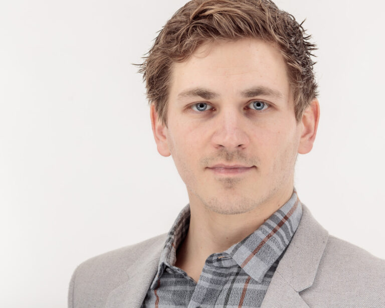 A man with brown hair and blue eyes wearing a grey blazer over a plaid shirt, photographed against a white background.