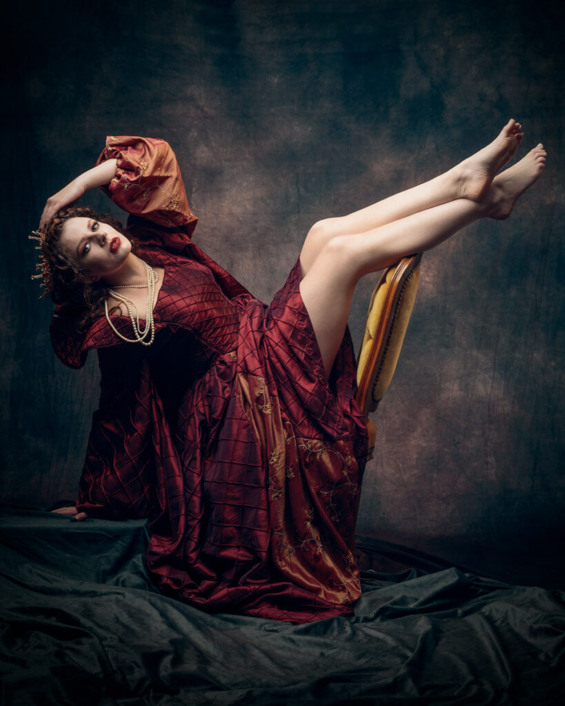 A woman in an ornate burgundy robe reclines on a chair with one leg raised, wearing pearl necklaces against a dark backdrop.
