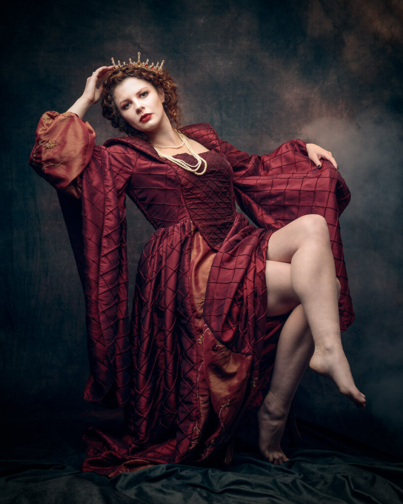 A woman in a burgundy quilted robe reclines against a dark backdrop, wearing a tiara and pearl necklace.