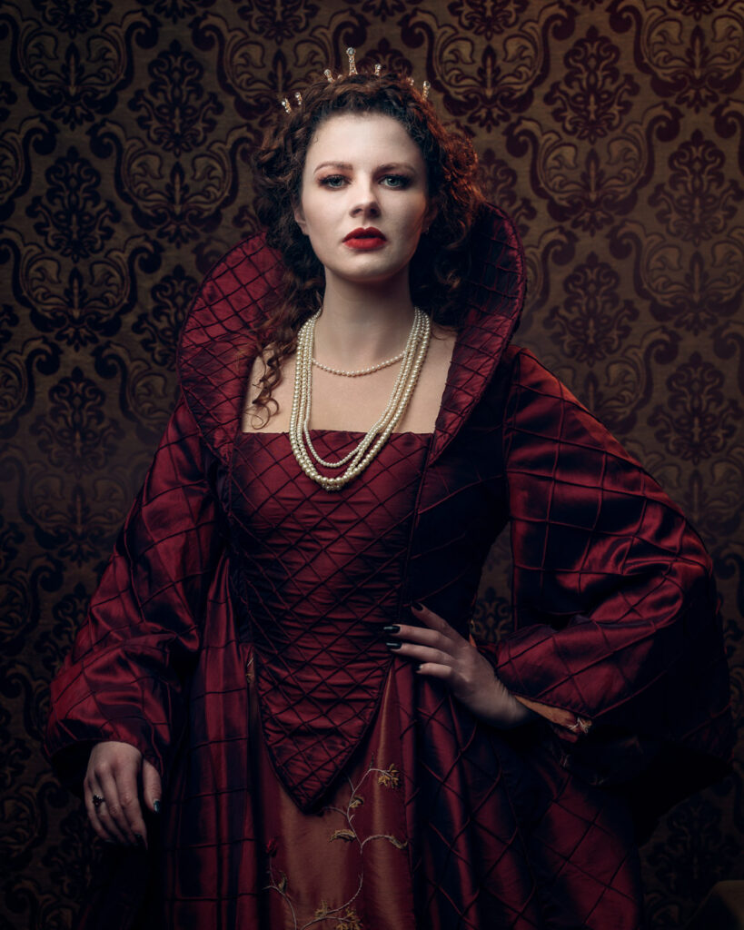 A woman in a red medieval gown with pearl necklace and tiara stands against a damask patterned background.