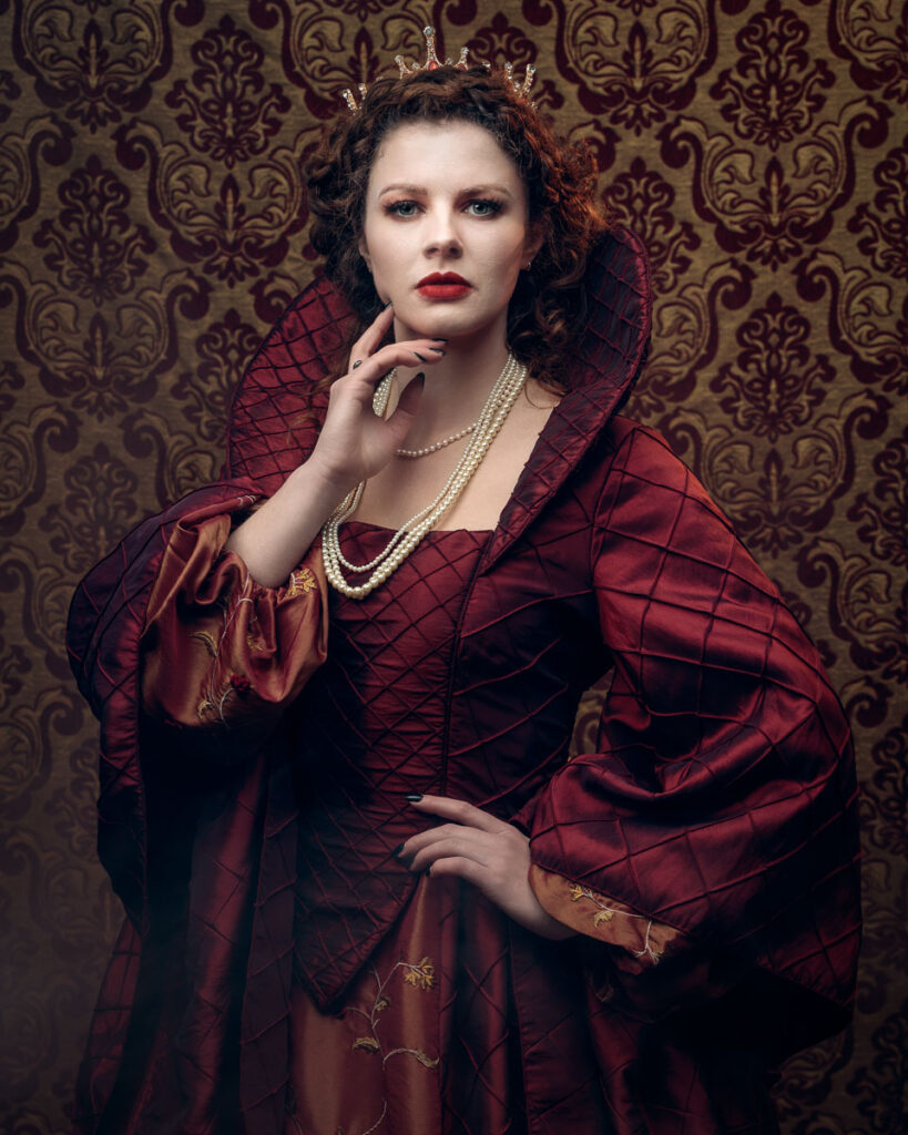 A woman in an ornate red gown and tiara stands against a damask patterned backdrop, wearing pearl necklaces and red lipstick.