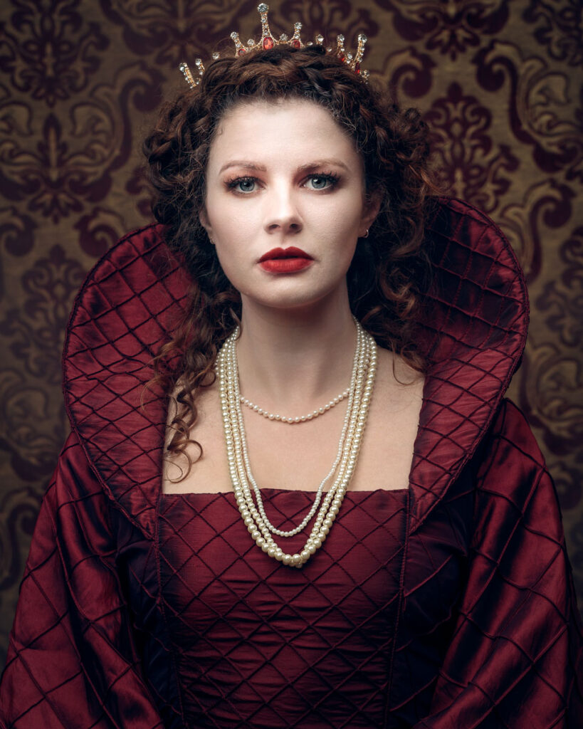 A woman wearing an ornate red gown, pearl necklaces and a jewelled crown sits against a damask wallpaper backdrop.