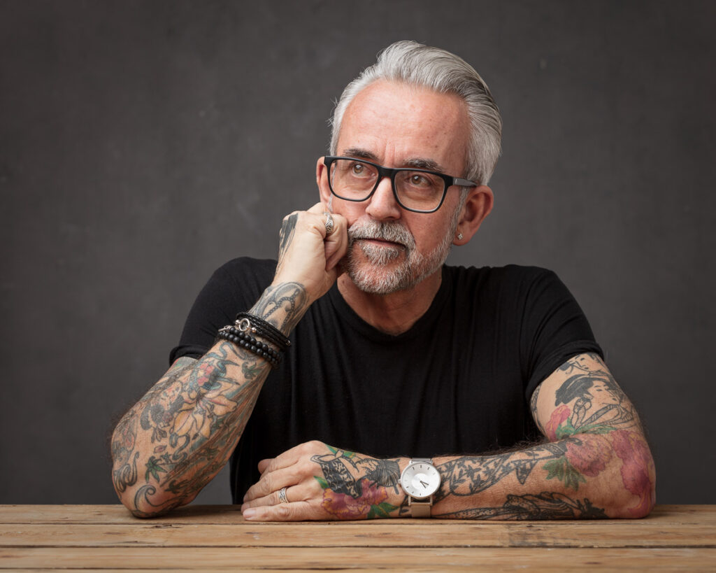 A man with grey hair and glasses sits at a wooden table, resting his chin on his hand. He has extensive tattoos on both arms and wears a black shirt and watch.