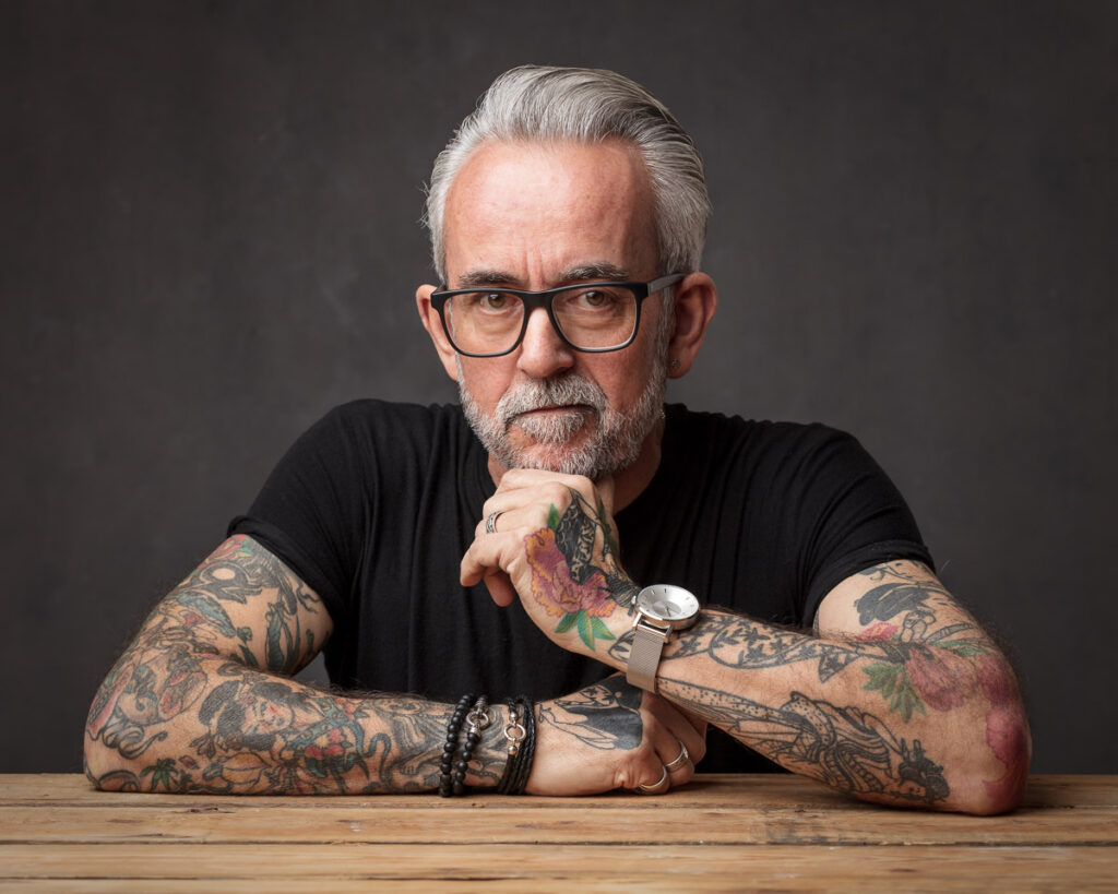 A man with grey hair and glasses, wearing a black shirt, sits at a wooden table with his arms crossed, displaying extensive tattoos on both arms.