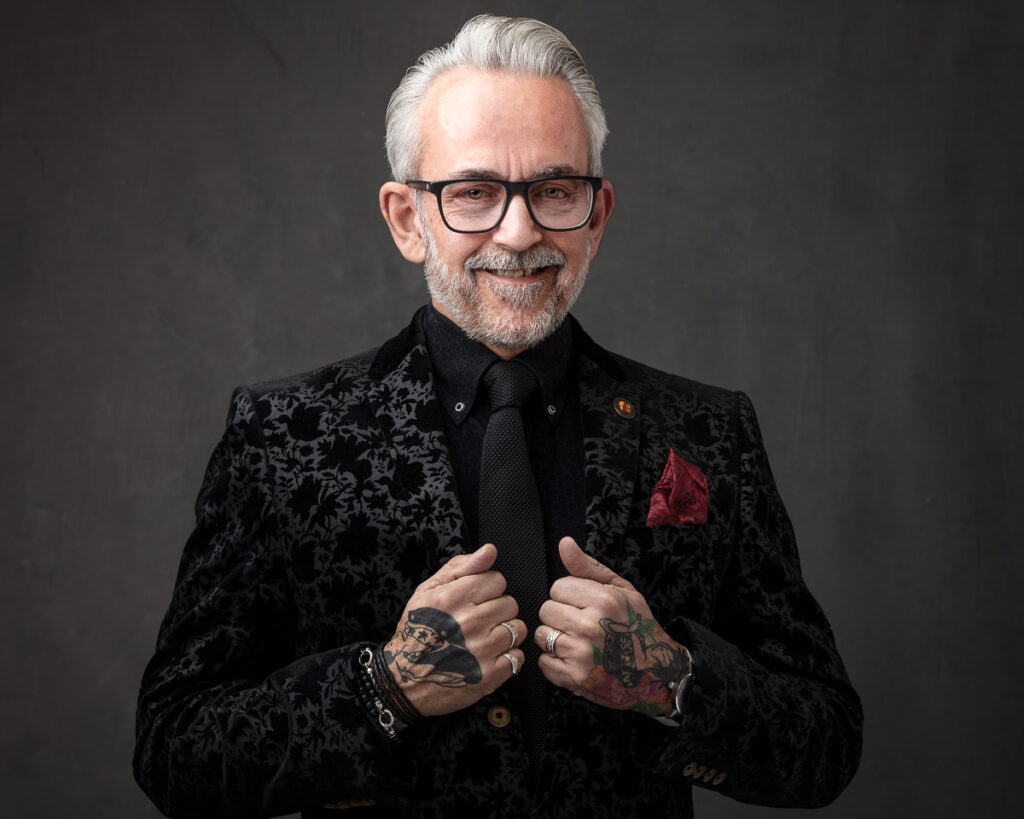 A man with white hair and glasses wears a patterned black jacket over a black shirt with tie. He has tattoos on his hands and is smiling at the camera against a grey background.