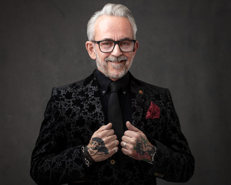 A man with white hair and glasses wears a patterned black jacket over a black shirt with tie. He has tattoos on his hands and is smiling at the camera against a grey background.