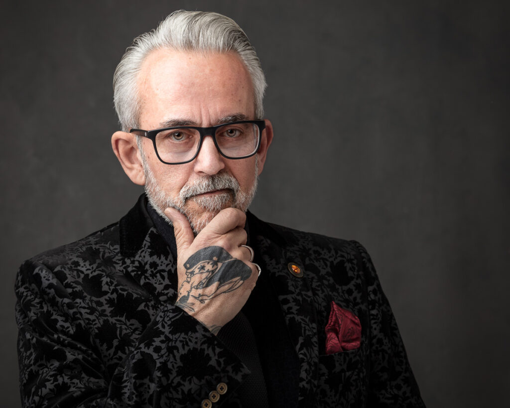An older man with white hair and glasses wears a black patterned jacket. He has a beard and tattoo on his hand, posed thoughtfully against a grey background.