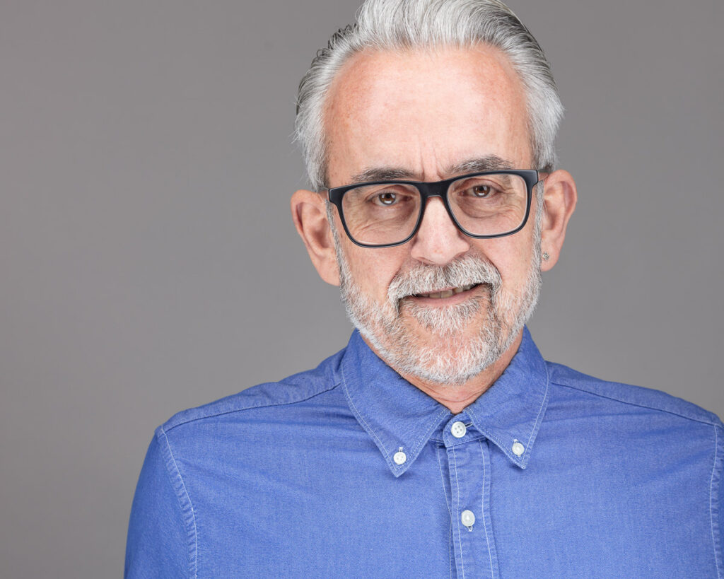 A man with white hair and glasses wearing a blue denim shirt, smiling at the camera against a grey background.