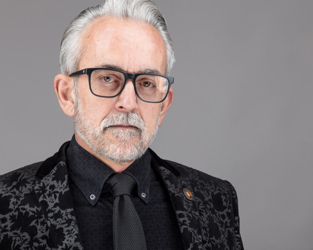 A man with white hair and glasses wears a black patterned jacket and tie against a grey background.