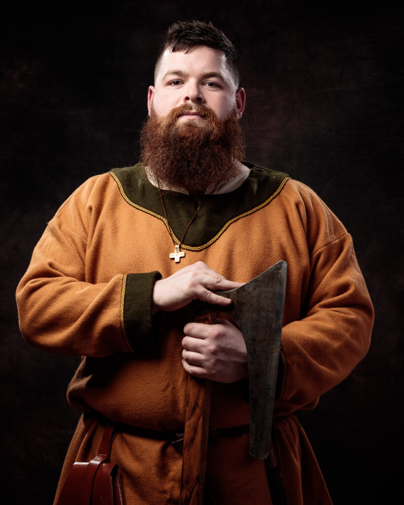 A bearded man in medieval-style clothing holds a dark cloth against a black background. He wears a tan tunic with green trim and a cross necklace.