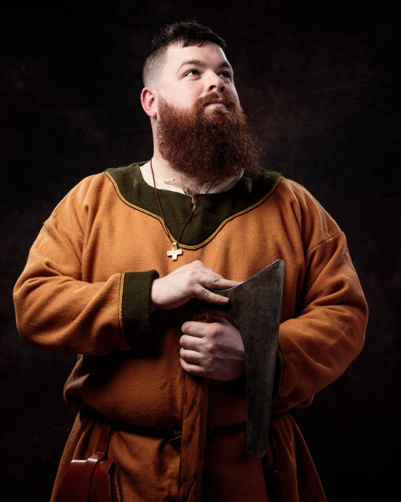 A bearded man in medieval-style brown tunic holds a dark book, wearing a cross necklace against a black background.