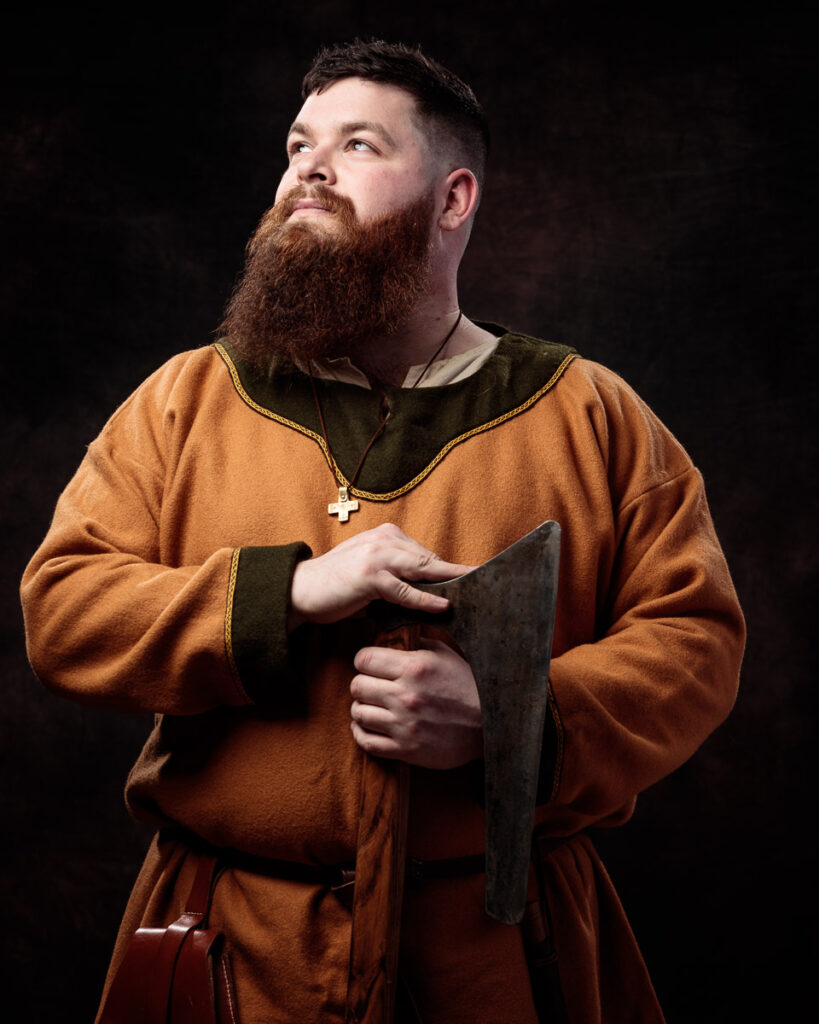 A bearded man in medieval-style brown tunic holds an axe, wearing a cross necklace against a dark background.