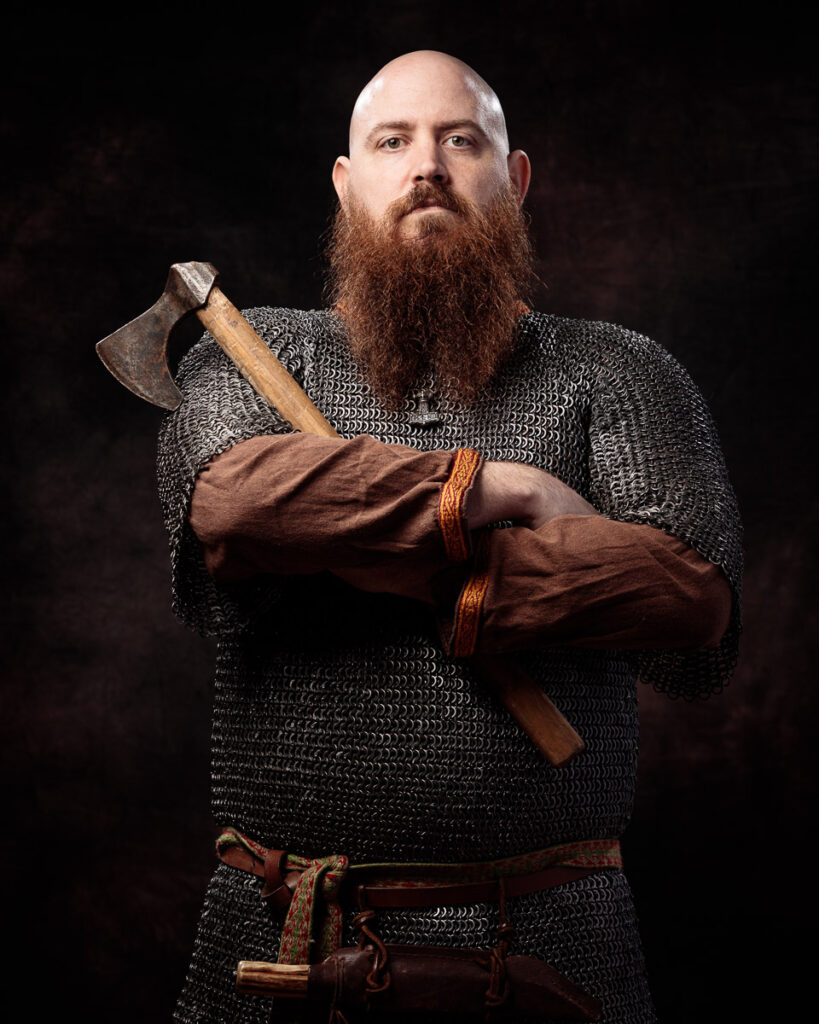 A bearded man in chainmail armour holds an axe across his chest against a dark background.