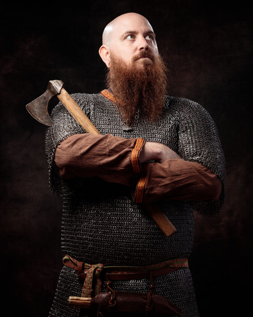 A bearded man in Viking-style chainmail armour holds an axe across his chest against a black background.