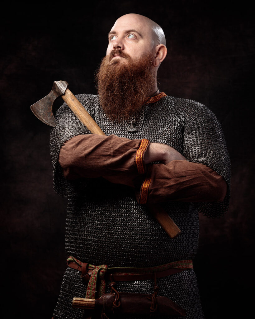 A bearded man in chainmail armour holds an axe across his chest against a dark background.