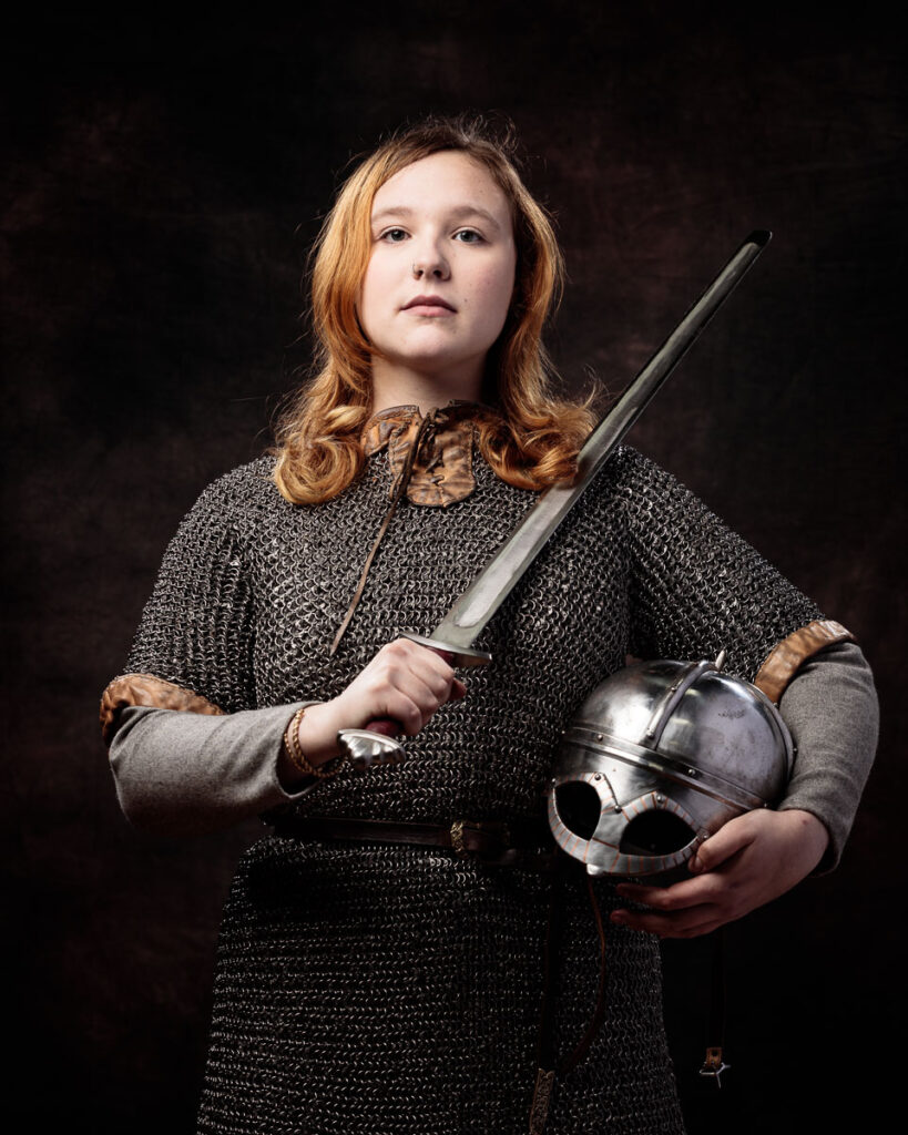 A young person with reddish-brown hair wearing chainmail armour holds a sword and metal helmet against a dark background.