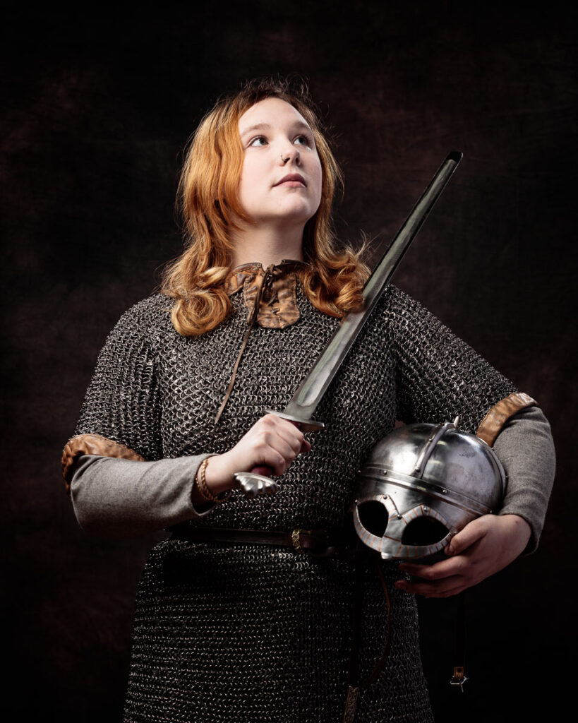 A young woman with auburn hair wearing chainmail and holding a sword and helmet, gazing upward against a dark background.