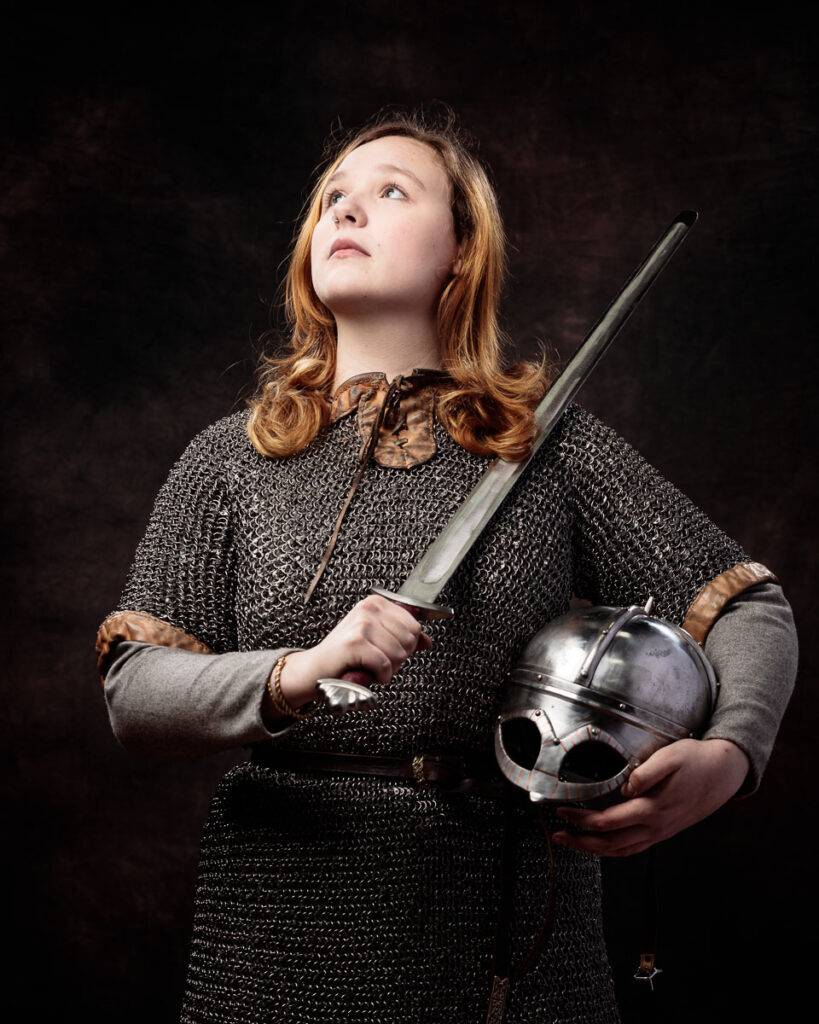 A young woman with reddish-brown hair wears chainmail armour and holds a sword and helmet against a dark background.