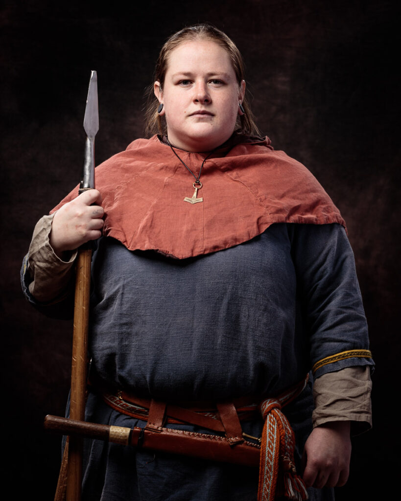 A young person in Viking-era costume holds a spear and axe against a dark background.