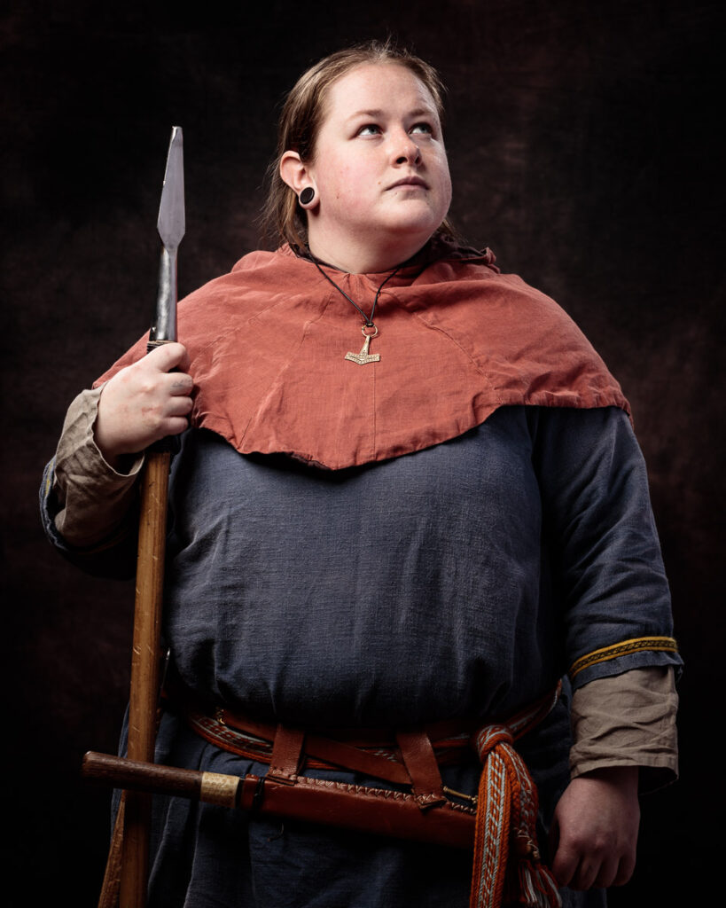 A person in Viking-era costume holds a spear, wearing a dark tunic with a rust-coloured cape and a Thor's hammer pendant.
