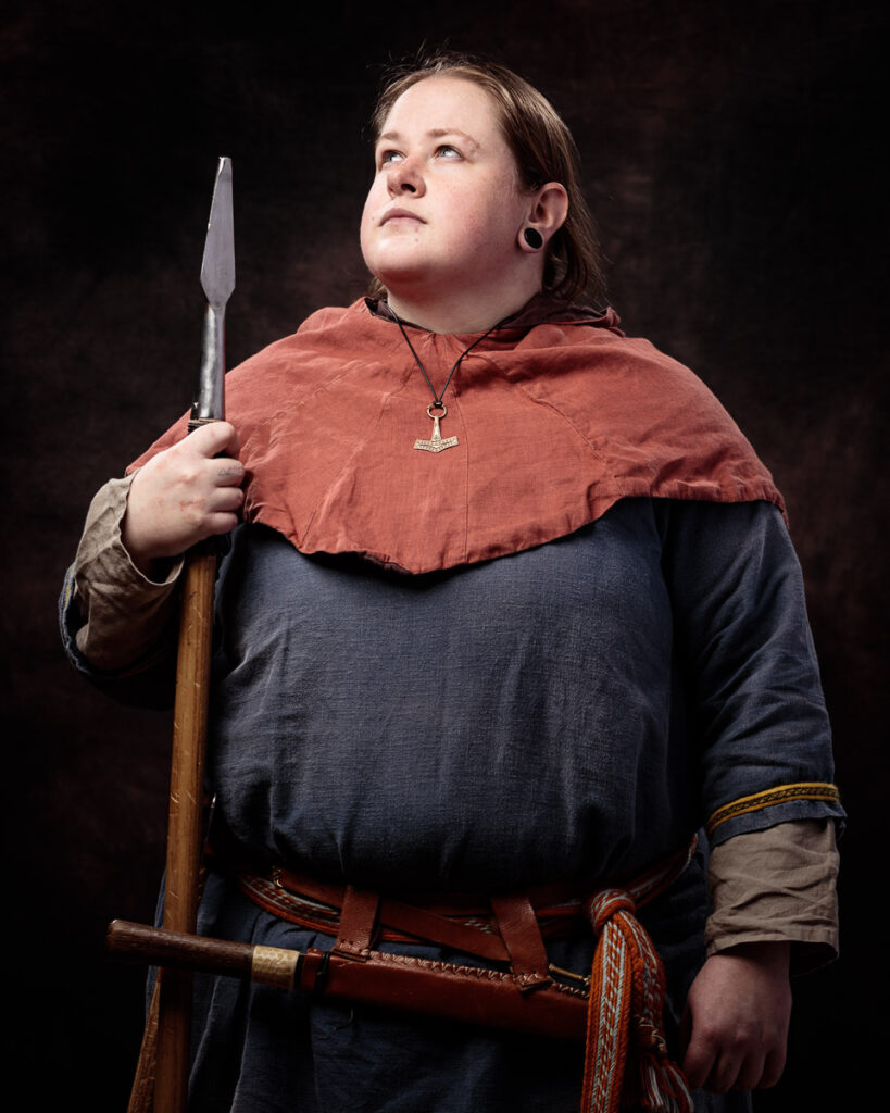 A person in Viking-era clothing holds a spear, wearing a dark tunic with a rust-colored cape and a pendant necklace, against a dark background.