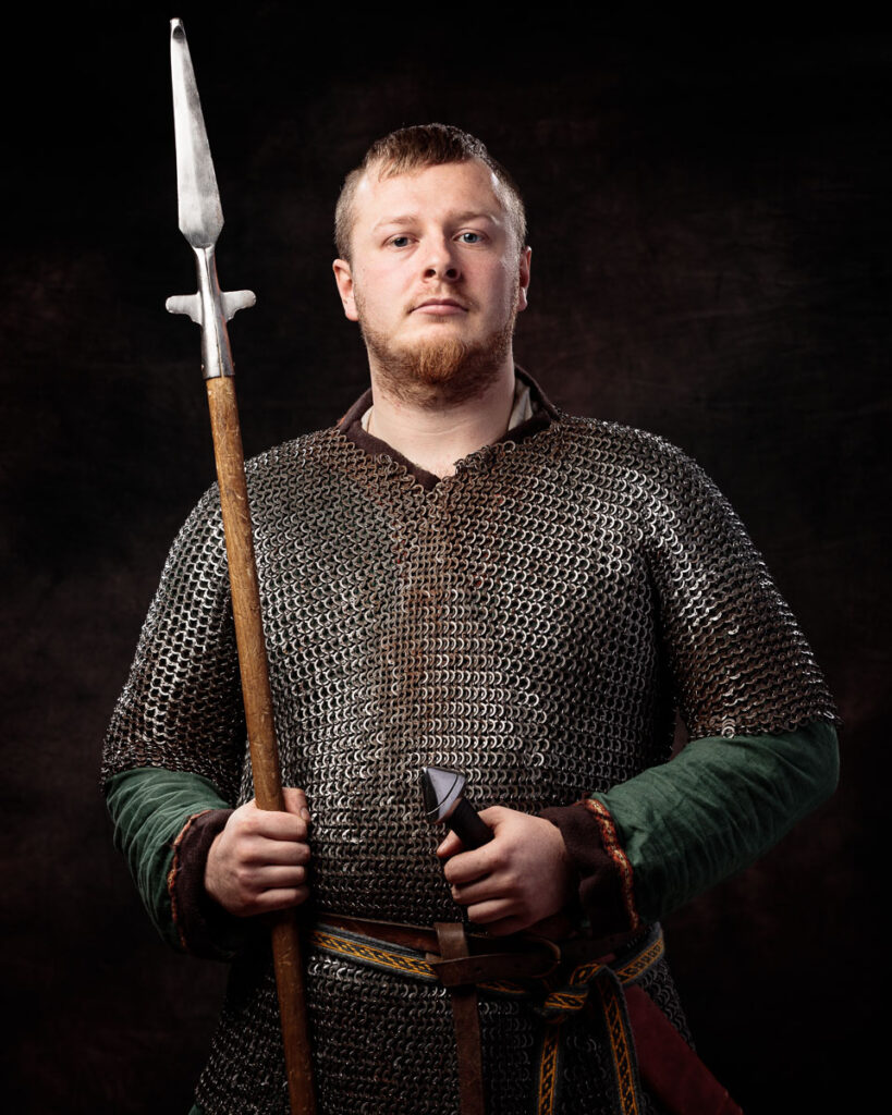 A bearded man in medieval chainmail armour holds a spear, posed against a black background.