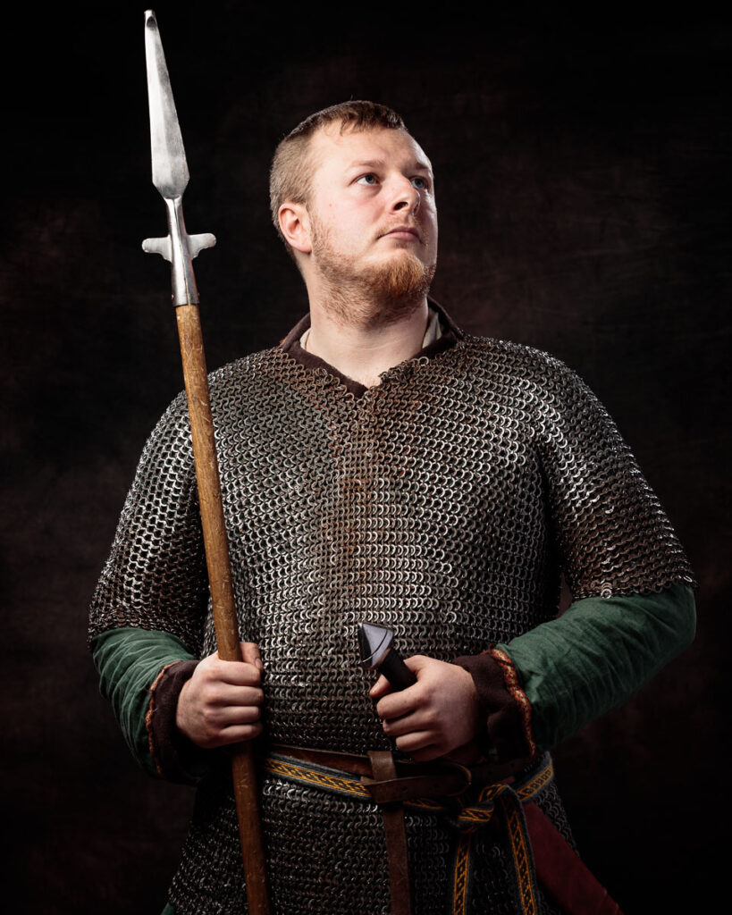 A bearded man in medieval chainmail armour holds a spear, gazing upward against a dark background.