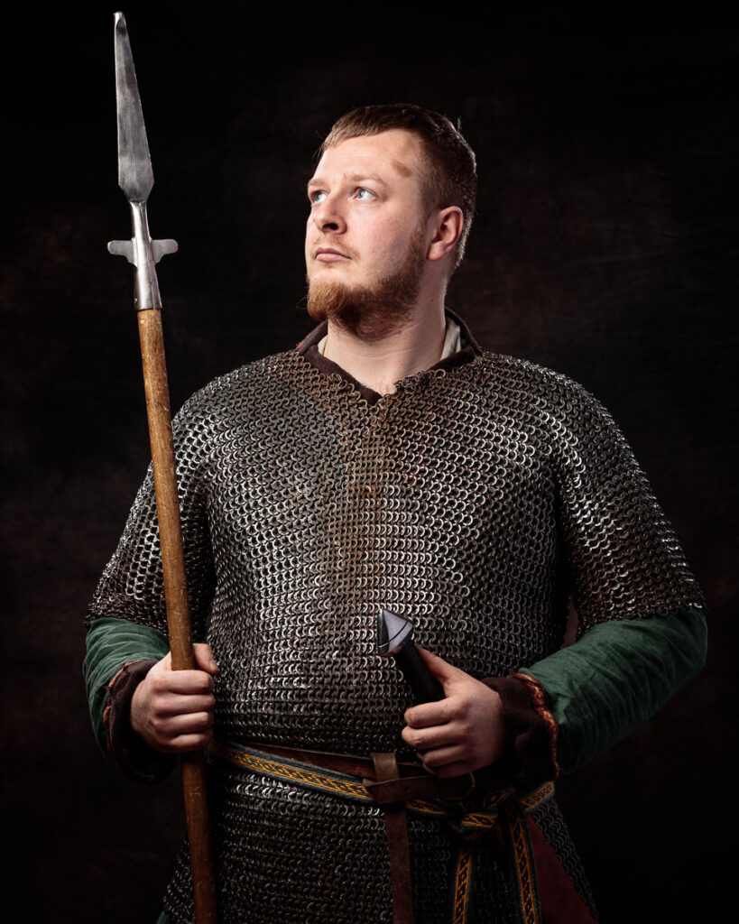 A bearded man in medieval chainmail armour holds a spear with a wooden shaft, set against a black background.