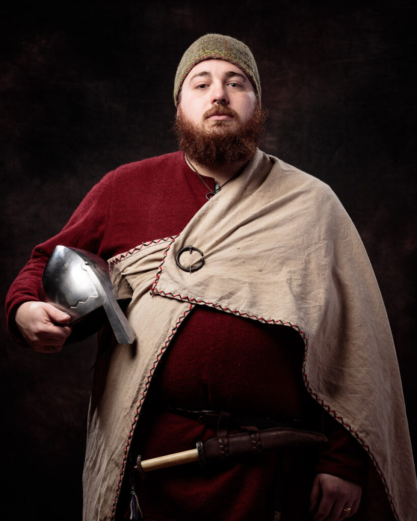 A bearded man in medieval clothing holds a helmet and stands against a dark background.