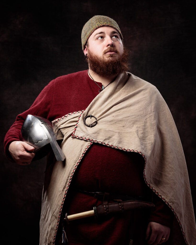 A bearded man in medieval clothing holds a helmet and axe against a dark background, looking upward thoughtfully.