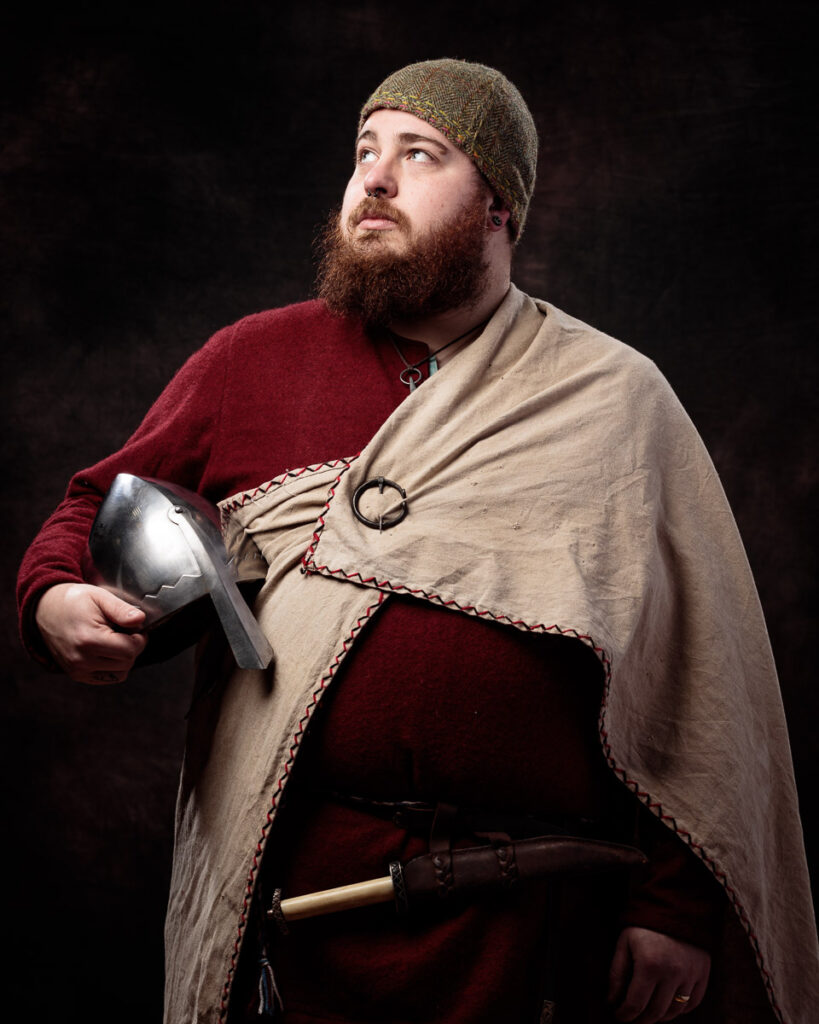 A bearded man in medieval costume holds a helmet, wearing a red tunic, beige cloak and wool hat against a dark background.