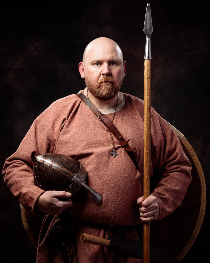 A bearded man in period costume holds a spear and shield against a dark background.