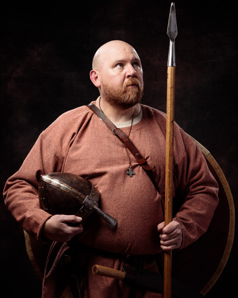 A bearded man in medieval clothing holds a spear and helmet against a dark background.