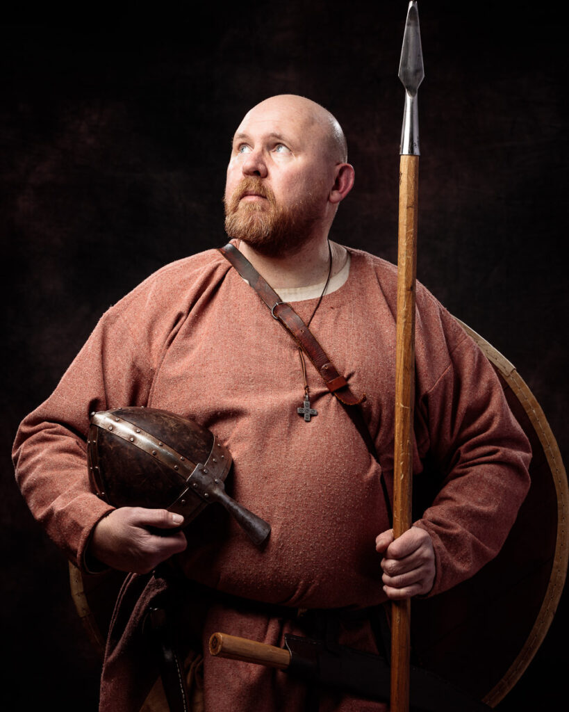 A bearded man in a rust-coloured tunic holds a spear and helmet against a dark background, dressed as a Viking warrior.