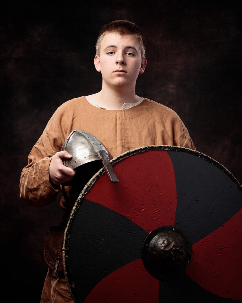 A young boy in a tan tunic holds a metal helmet and a red and black shield against a dark background.