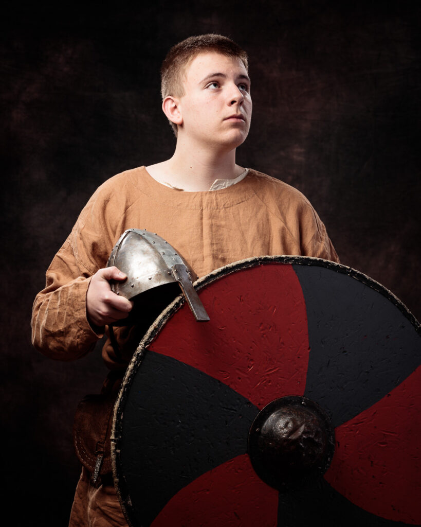 Young boy in brown tunic holding a metal helmet and red and black shield, looking upward against a dark background.