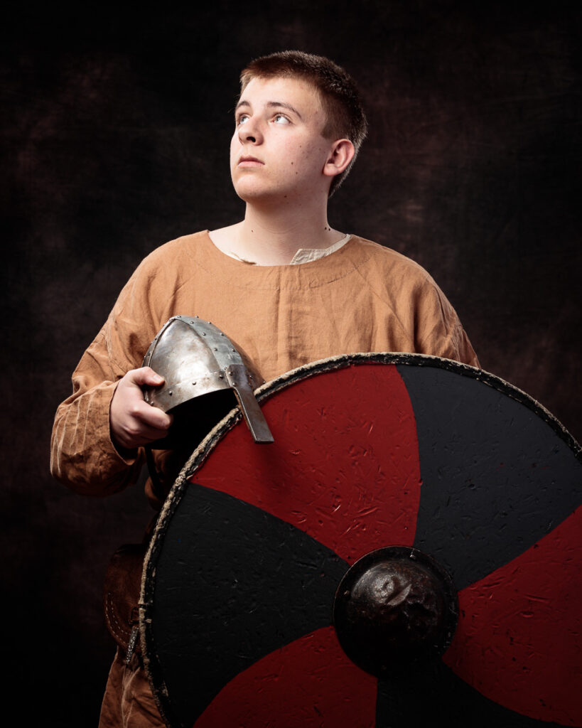 A young person in a tan tunic holds a metal helmet and a red and black shield, gazing upward against a dark background.