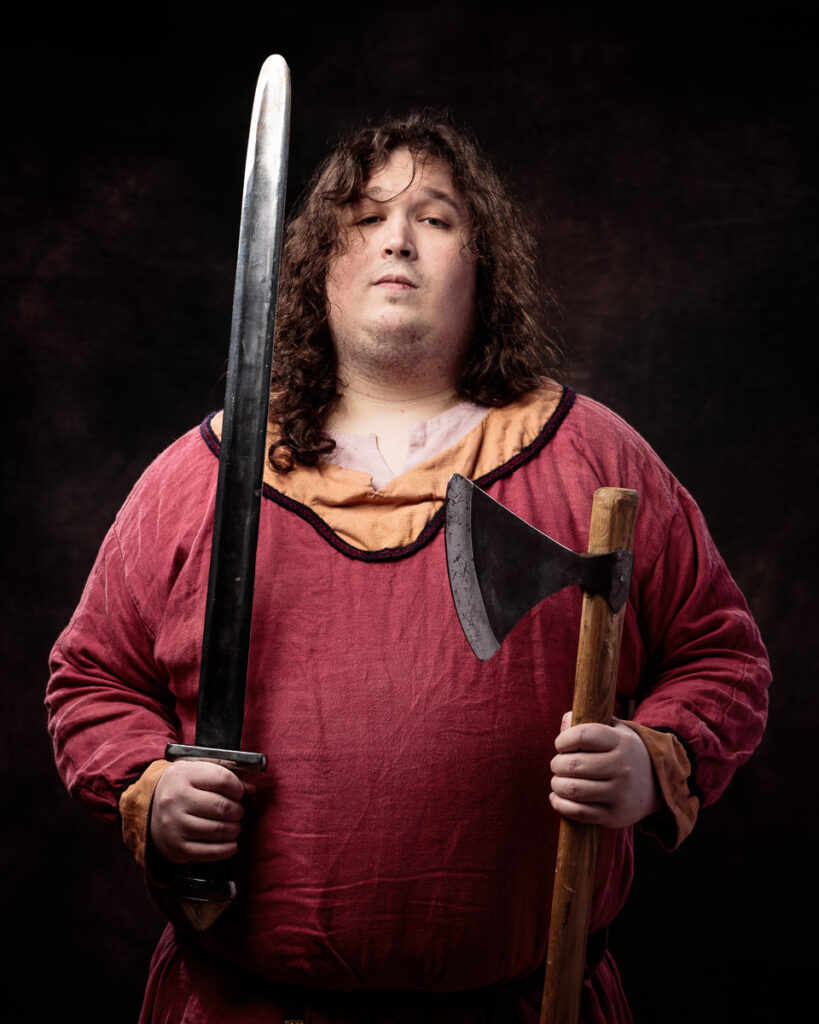 A man with long curly brown hair wearing a burgundy tunic holds a sword in one hand and an axe in the other against a dark background.