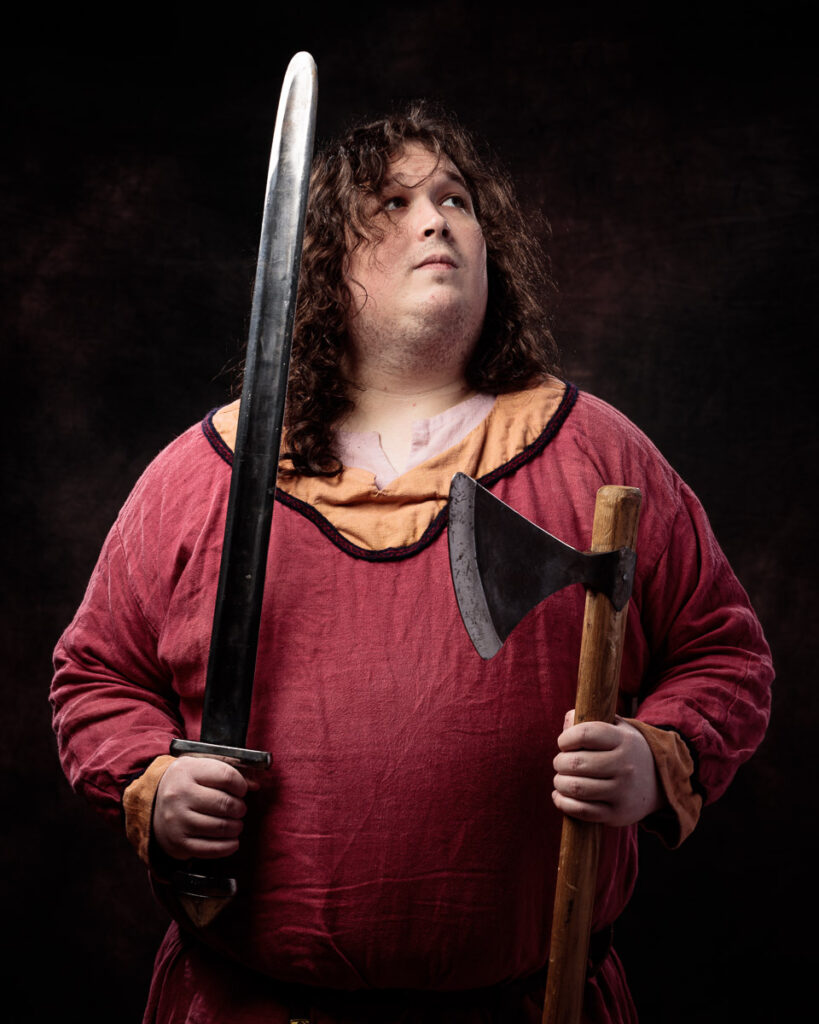 A person with long curly brown hair wearing a burgundy tunic holds a sword and axe against a dark background.