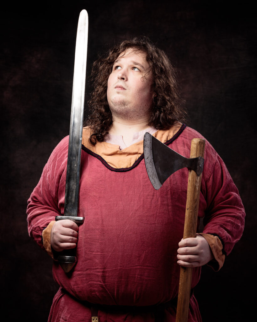 A man with long curly dark hair wearing a burgundy tunic holds a sword and axe against a black background.
