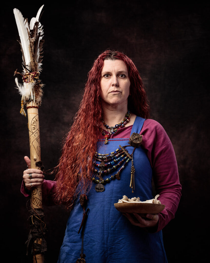 A person with long burgundy hair holds a decorative staff with feathers and wears medieval-style clothing against a dark background.